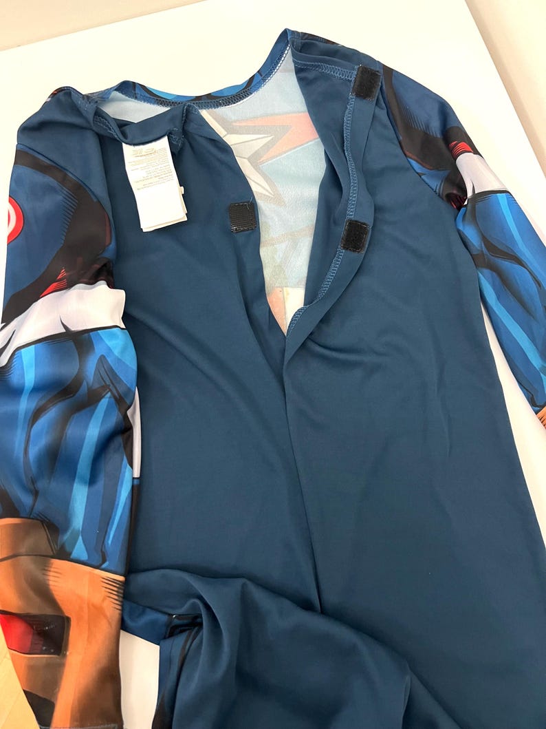 May include: A blue superhero costume with long sleeves and a mock neck. The costume has a graphic design on the sleeves and chest, with hook and loop fasteners down the front. A white tag is attached to the costume.
