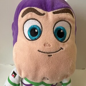 Pode incluir: Um brinquedo de pel&uacute;cia do personagem Buzz Lightyear da franquia Toy Story. O brinquedo tem uma cabe&ccedil;a roxa, um rosto bege e olhos azuis grandes. Ele usa um traje espacial branco e verde com um emblema preto e vermelho.