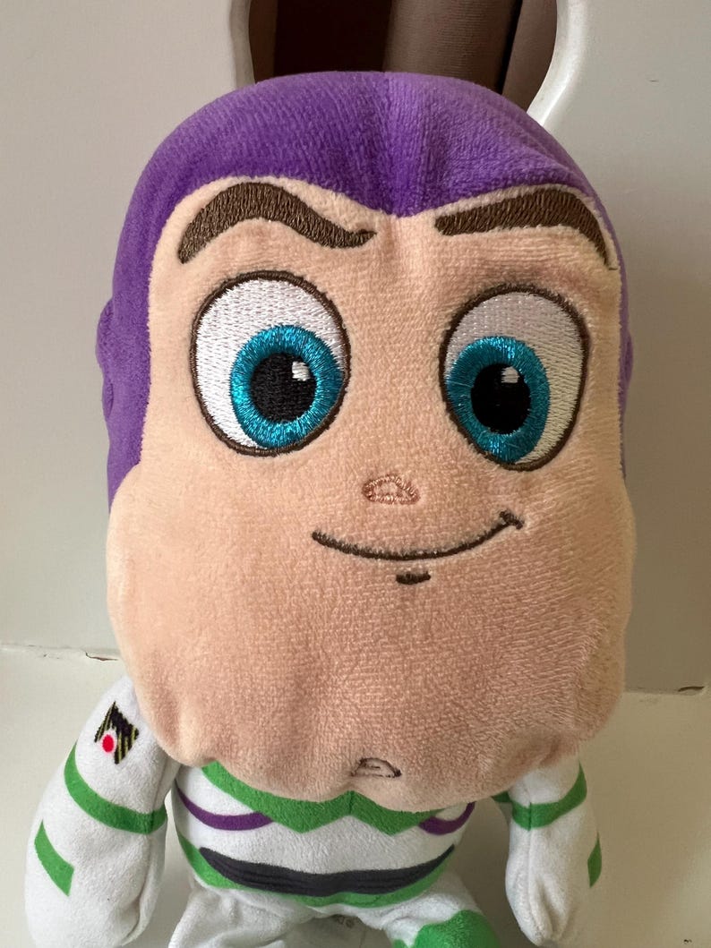 Pode incluir: Um brinquedo de pel&uacute;cia do personagem Buzz Lightyear da franquia Toy Story. O brinquedo tem uma cabe&ccedil;a roxa, um rosto bege e olhos azuis grandes. Ele usa um traje espacial branco e verde com um emblema preto e vermelho.