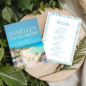 May include: Two printed cards on a wooden tray surrounded by greenery. One card reads "DANIELLE'S BACHELORETTE" with a beach scene. The other card has a teal border and the word "details" with a schedule. The background includes leaves and dried plants.