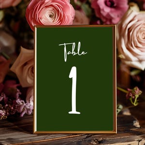 May include: A framed table number sign with a dark green background and gold trim. The sign displays the word "Table" and the number "1" in white script. The background includes pink and purple flowers.