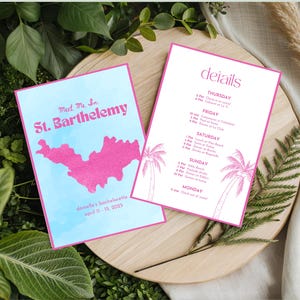 May include: Two pink and blue event cards with the text "Meet Me In St. Barthelemy" and "details". The cards are on a wooden tray surrounded by greenery and include a map of St. Barthelemy and palm tree illustrations.