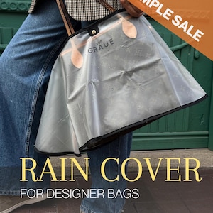 Pode incluir: Uma capa de chuva de plástico transparente e flexível para bolsa, com acabamento preto, alças em tom de couro castanho e o texto 'von GRAUE' impresso na frente. A superfície apresenta pequenas gotas de água para proteção da bolsa.