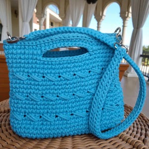 May include: A turquoise crochet handbag with a handle and a detachable shoulder strap. The bag features a textured pattern and silver-tone hardware. The bag is approximately 23 cm wide and 20 cm tall.