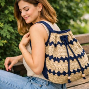 Op de afbeelding: Een beige en marineblauwe gehaakte rugzak met een trekkoordsluiting. De tas heeft een geometrisch patroon in marineblauw en een marineblauwe band. De tas is gemaakt van zacht, getextureerd garen en heeft houten kraalaccenten.