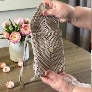 Op de afbeelding: Een beige gehaakte crossbody tas met een chevronpatroon. De tas heeft een klepsluiting met een magneetsluiting en een lange riem. De tas is open, waardoor de binnenkant zichtbaar is. Een vaas met roze bloemen staat op de achtergrond.