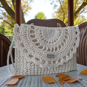 May include: A cream-colored, crocheted handbag with a flap closure. The flap features a decorative, semi-circular pattern. The bag has a long strap and a small, rectangular metal tag. The bag is sitting on a table with fallen leaves.