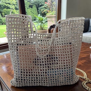 Op de afbeelding: Een witte, grofmazige gehaakte tote bag met een vierkant rasterpatroon, twee geïntegreerde handvatten en een goudkleurige metalen kettingriem aan de zijkant. De tas heeft een rechthoekig silhouet en een semi-transparante constructie.
