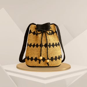 May include: A handmade crochet bucket bag in a golden yellow color with black zigzag patterns. The bag has black straps and a drawstring closure. Two wooden beads adorn the drawstring. The bag is sitting on a gold-colored round platform.