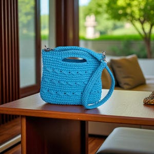 以下が含まれることがあります： ターコイズブルーのかぎ針編みハンドバッグ。上部のハンドルと取り外し可能なショルダーストラップが付いています。バッグは、テクスチャードステッチパターンとシルバーカラーのハードウェアが特徴です。バッグは木製のテーブルの上にあります。