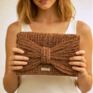 May include: A brown, rectangular clutch bag with a large bow detail. The bag is made of a textured, woven material and features a small, rectangular metal tag. The bag is held up by a person.