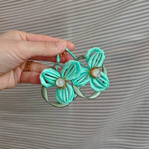 Könnte beinhalten: Ein Paar Hängeohrringe mit leuchtend türkisfarbenen, stoffumwickelten Blütenblättern in Blumenform. Jeder Ohrring hat eine zentrale, schillernde weiße Perle, die von kleinen goldfarbenen Metallspitzen umgeben ist und an einem runden Rahmen befestigt ist.