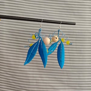 Puede incluir: Un par de pendientes colgantes con hojas de alambre azul brillante, pequeñas cuentas esféricas amarillas y cuentas redondas más grandes de color melocotón pálido. Los pendientes cuelgan de ganchos de metal plateado y tienen un diseño botánico.