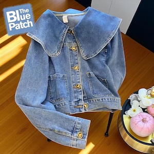 Könnte beinhalten: Hellblaue Jeansjacke mit großem, rundem Kragen. Die Jacke hat goldfarbene Knöpfe und zwei Vordertaschen. Der Text "The Blue Patch" befindet sich in der oberen linken Ecke.