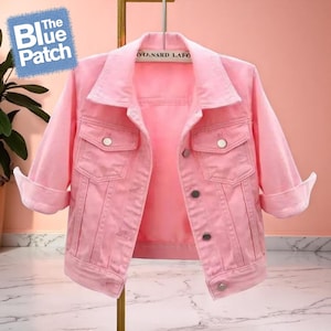 Könnte beinhalten: Hellrosa Jeansjacke mit klassischem Kragen, Knopfleiste und zwei Brusttaschen. Die Ärmel sind hochgekrempelt. Die Jacke hängt an einem goldenen Kleiderbügel. Das Logo The Blue Patch befindet sich in der oberen linken Ecke.