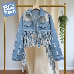 Puede incluir: Chaqueta vaquera azul claro con detalles desgastados y flecos. La chaqueta tiene cuello clásico, cierre de botones y dos bolsillos en el pecho. El logo The Blue Patch es visible.