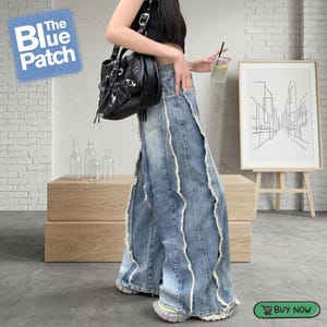Op de afbeelding: Lichtblauwe jeans met golvende, witte distressed details. De jeans heeft een wijde pijp en een hoge taille. De afbeelding toont ook een zwarte handtas en een ingelijste kunstdruk.
