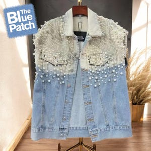 Könnte beinhalten: Hellblaue Jeansjacke mit Perlen- und Strassverzierungen. Die Jacke hat einen klassischen Kragen, eine Knopfleiste und zwei Brusttaschen. Das Logo The Blue Patch ist oben links sichtbar.