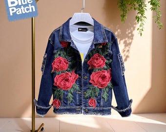 May include: A dark blue denim jacket with large red rose embroidery. The jacket has a classic collar, button closure, and frayed edges. The logo "The Blue Patch" is in the upper left corner. The jacket is hanging on a gold-colored rack.