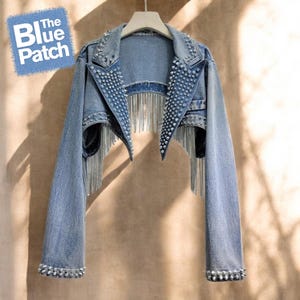 Puede incluir: Chaqueta vaquera azul claro con tachuelas plateadas y flecos de cadena. La chaqueta presenta un diseño corto con mangas largas y está adornada con tachuelas plateadas a lo largo del cuello, los hombros y los puños. El logotipo de The Blue Patch está arriba a la izquierda.
