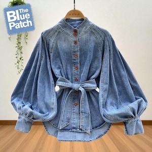 May include: Light blue denim shirt with a mandarin collar, button-down front, and a waist tie. The shirt features long, voluminous sleeves with buttoned cuffs. The Blue Patch logo is visible in the upper left corner.