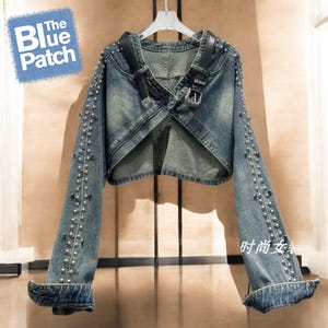 Könnte beinhalten: Eine kurze Jeansjacke in verwaschenem Blau, verziert mit silberfarbenen Nieten an den Ärmeln und schwarzen Lederriemendetails. Die Jacke hat ein einzigartiges offenes Frontdesign und umgeschlagene Manschetten. Der Text "The Blue Patch" befindet sich in einem blauen Patch.
