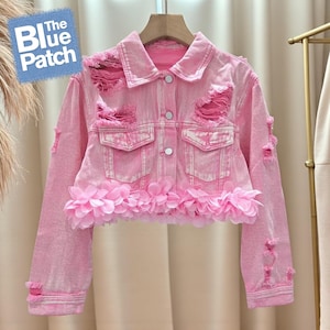 May include: Pale pink denim jacket with ripped details and a floral trim. The jacket features a classic collar, button closure, and two front pockets. The Blue Patch logo is visible in the upper left corner.