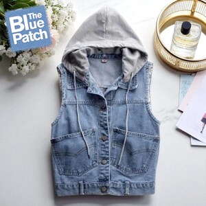 Puede incluir: Un chaleco vaquero azul claro con capucha gris. El chaleco tiene dos bolsillos delanteros, cierre de botones y cordones blancos. El logotipo de Blue Patch es visible. El chaleco es sin mangas.