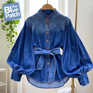 Puede incluir: Una camisa vaquera azul con cuello alto y mangas largas y voluminosas. La camisa tiene una botonadura frontal, una cinturón en la cintura y el texto "The Blue Patch" en un cuadrado azul.