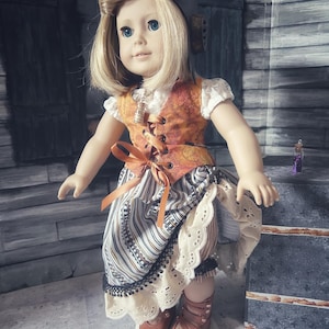 May include: A blonde doll wearing a brown aviator cap, a white short-sleeved blouse, and an orange patterned vest with brown ribbon lacing. The doll wears a striped skirt with a cream-colored eyelet lace hem and brown strappy leather-look shoes.