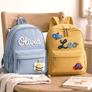 Puede incluir: Dos mochilas de pana, una azul claro y otra amarilla, con nombres bordados y parches. La mochila azul muestra el nombre Olivia y un parche de abeja. La mochila amarilla muestra el nombre Leo, un avión azul y un parche de coche rojo.