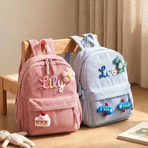 Peut inclure: Deux sacs à dos en velours côtelé, un rose et un bleu clair, avec les prénoms Lily et Leo brodés. Le sac rose porte une fleur et un gâteau en peluche, tandis que le bleu présente un dinosaure vert et deux avions bleus avec des tirettes tressées.