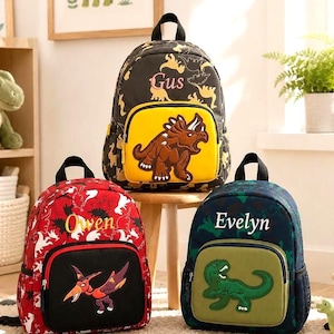 Puede incluir: Tres mochilas con motivos de dinosaurios y nombres personalizados. La bolsa negra superior muestra 'Gus' con un triceratops. La bolsa roja muestra 'Owen' con un pterodáctilo. La bolsa azul oscuro presenta 'Evelyn' con un tiranosaurio rex verde.