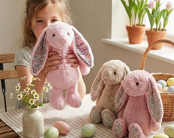 May include: A child holds a pink plush bunny with floral print ears. Two other bunnies, one beige and one pink, sit on a checkered tablecloth. Easter eggs in pastel colors and a basket are also present. A vase of flowers and potted tulips complete the scene.