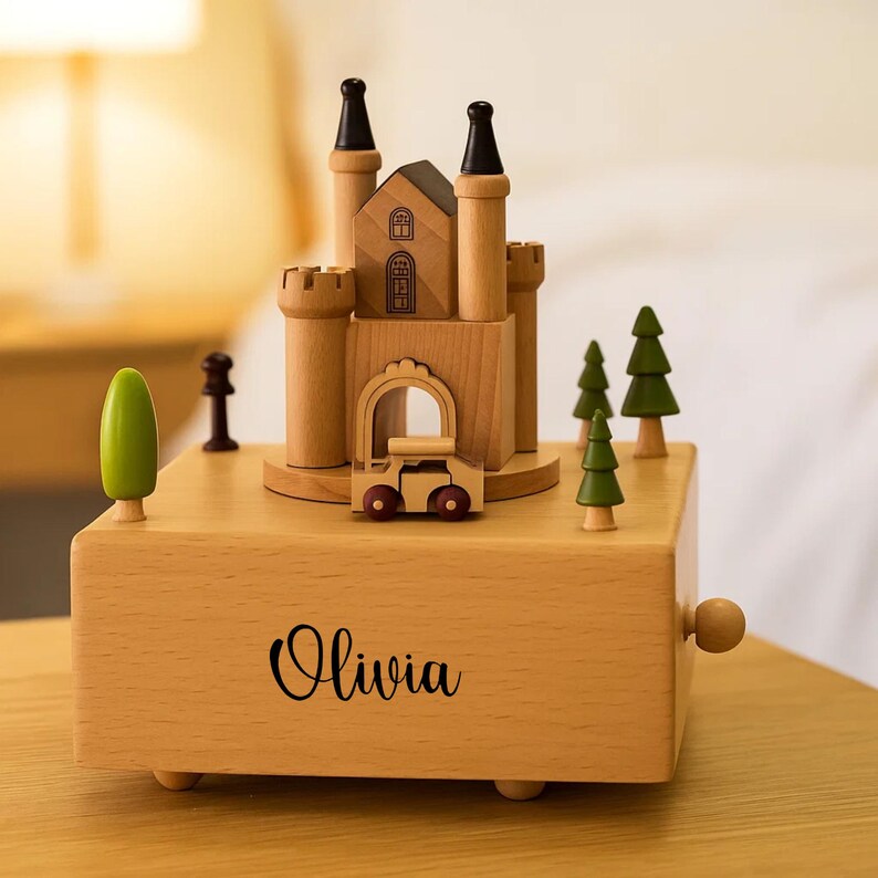 May include: A wooden music box in the shape of a castle, with a small car and green trees. The light brown box has the name "Olivia" printed on the front. The castle has black accents on the towers.