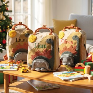 May include: Three beige canvas backpacks featuring textured, fuzzy dinosaur appliqu&eacute;s in green and rust colors. Each bag has a top handle, a front pocket, and a yellow sun patch. Two bags are personalized with the name Liam and one with Olivia.