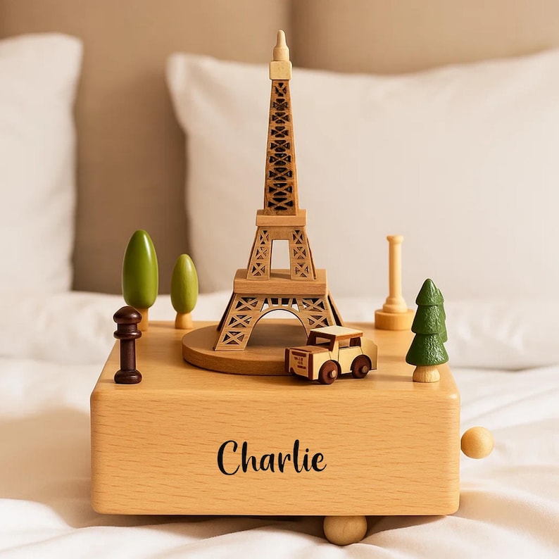 May include: Wooden music box with a miniature Eiffel Tower, a small car, and other decorative elements. The box is light-coloured with the name "Charlie" printed on the front. Includes a small green tree and other wooden accents.
