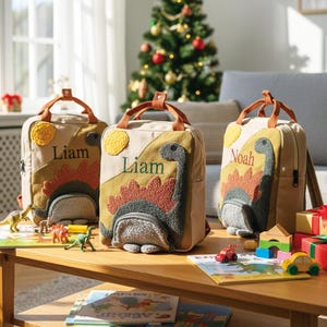 May include: Three beige canvas backpacks featuring textured dinosaur appliques in green, orange, and gray. Each bag displays a yellow sun and a personalized name, Liam or Noah, embroidered in dark green or red thread with brown top handles.