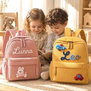 Könnte beinhalten: Ein rosafarbener Kordrucksack mit dem Namen Lunna und einem weißen Hasenaufnäher sowie ein gelber Kordrucksack mit dem Namen Leo, einem blauen Flugzeugaufnäher und einem roten Autoaufnäher. Beide Rucksäcke haben Vordertaschen mit Reißverschlüssen.