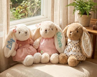 May include: Three plush bunny rabbits with long ears, each wearing a dress. One is in a pink gingham dress, another in a pink dress, and the third in a polka dot dress. The ears are embroidered with names.