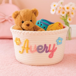 May include: A woven cream-colored basket with the name "Avery" in colorful stitched letters. The basket contains a brown teddy bear and some baby clothes. A yellow flower and a blue flower decorate the basket.