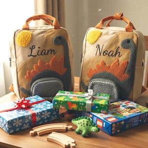 May include: Two beige canvas backpacks featuring embroidered dinosaur designs with textured orange and gray details. One bag is personalized with the name Liam and the other with Noah. Each backpack has top handles and a front pocket.