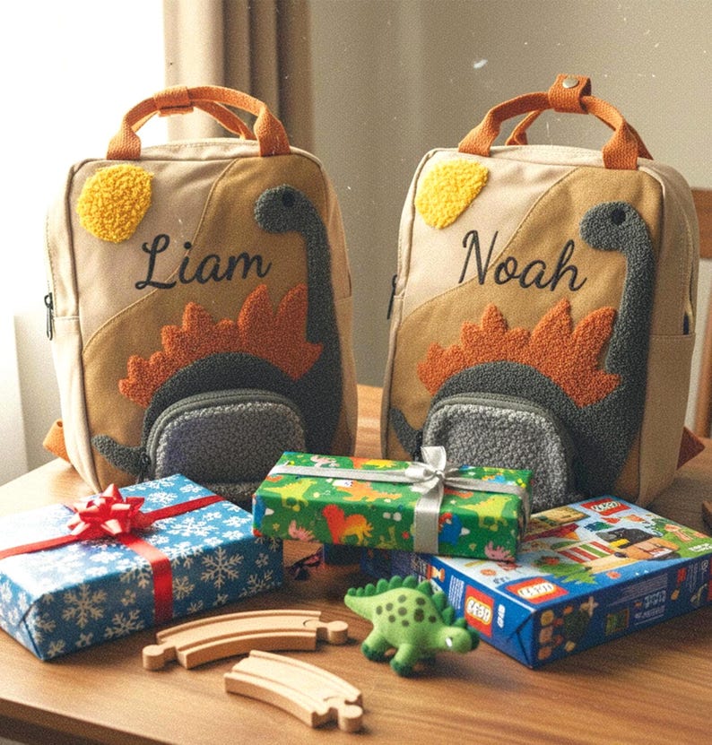May include: Two beige canvas backpacks featuring embroidered dinosaur designs with textured orange and gray details. One bag is personalized with the name Liam and the other with Noah. Each backpack has top handles and a front pocket.