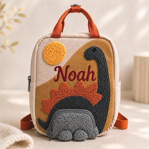 May include: A beige backpack featuring a textured orange sun, a dark grey dinosaur with orange spikes, and the name Noah embroidered in red. The bag has orange top handles, adjustable straps, and a small zippered pocket on the dinosaur's body.