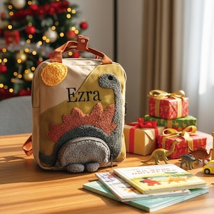 May include: A beige canvas backpack featuring a textured, dark gray and orange dinosaur appliqu&eacute; with a yellow sun. The name Ezra is embroidered in black script on the front. The bag has two brown top handles and a zippered front pocket.