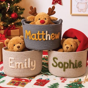 May include: Three woven rope baskets with personalized names: "Matthew" in yellow, "Emily" in white, and "Sophie" in green. Each basket contains a plush toy, including a reindeer and teddy bears. Christmas decor is in the background.