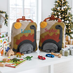 May include: Two beige canvas backpacks featuring a textured dark gray dinosaur appliqu&eacute; with a rust-colored spiky back, a yellow sun, and a front pocket. One backpack is embroidered with the name Liam in blue, and the other with Sophie in pink.