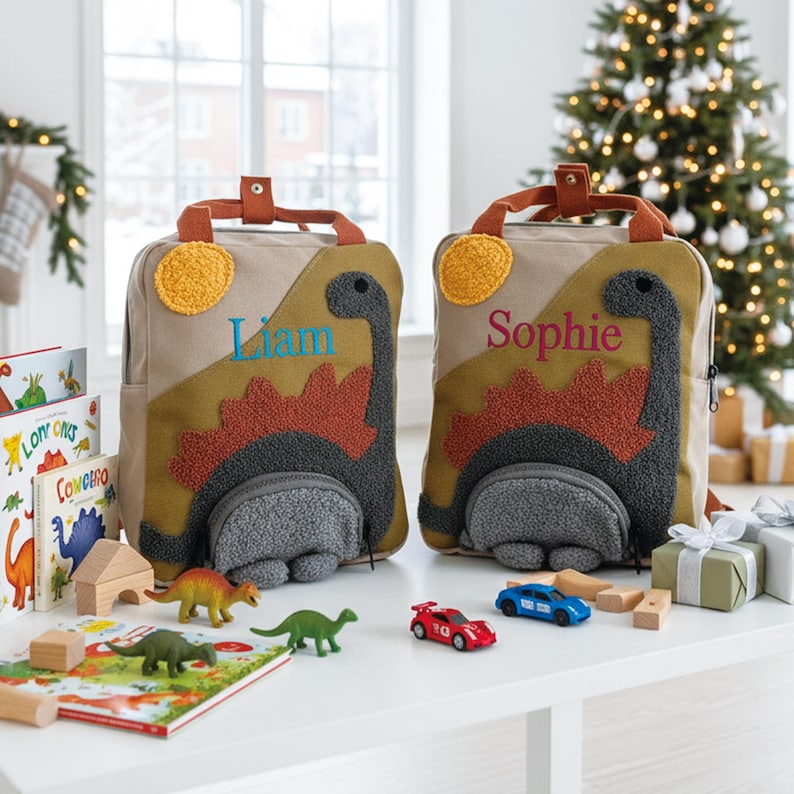 May include: Two beige canvas backpacks featuring a textured dark gray dinosaur appliqu&eacute; with a rust-colored spiky back, a yellow sun, and a front pocket. One backpack is embroidered with the name Liam in blue, and the other with Sophie in pink.