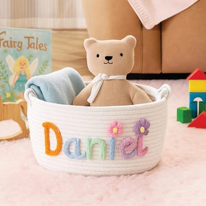May include: A white rope basket with the name "Daniel" in colorful letters and flower accents. Inside the basket is a tan teddy bear with a white ribbon and a blue blanket. A children's book and toy blocks are in the background.