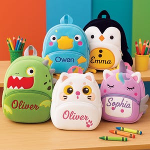 Puede incluir: Una colección de mochilas coloridas con forma de personajes. Los diseños incluyen un dinosaurio verde, un pato azul, un gato blanco, un pingüino blanco y negro y un unicornio morado. Cada mochila tiene un nombre bordado.